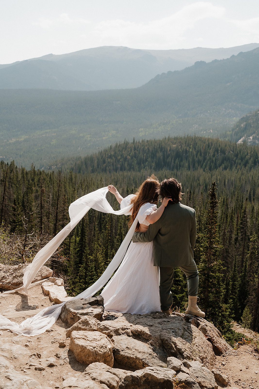 Rocky Mountain National Park elopement couple standing on rocky overlook with veil blowing in the wind above the Colorado forest