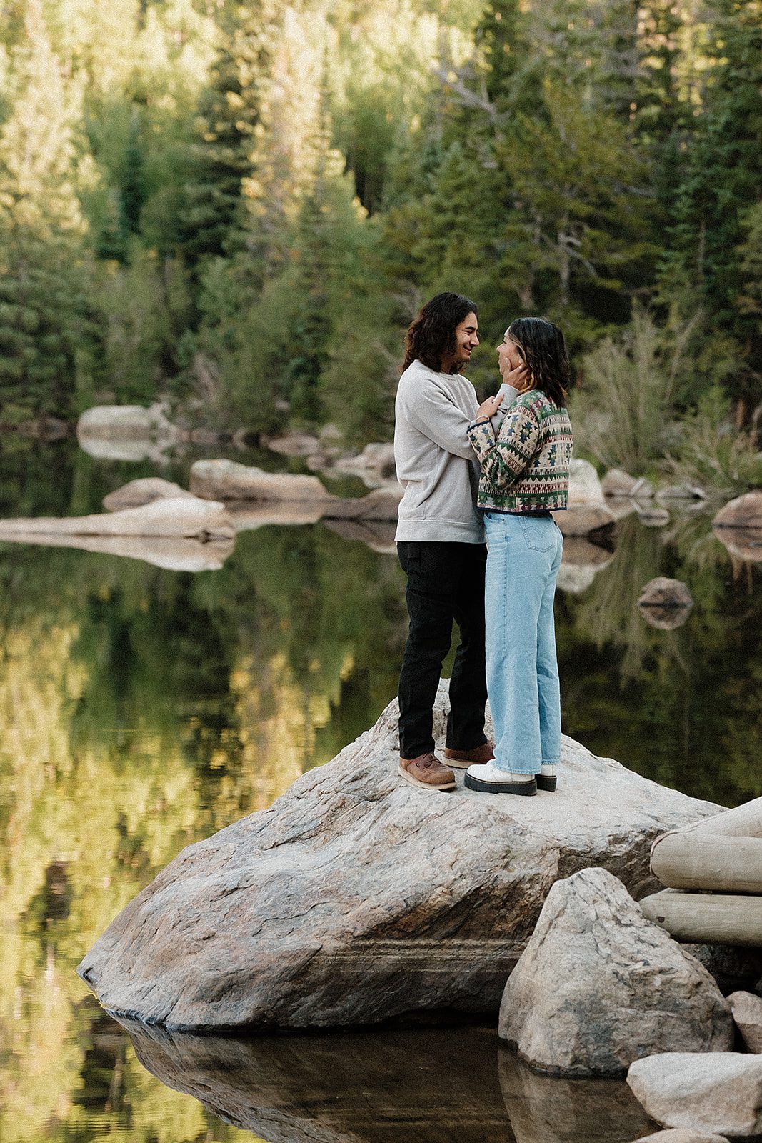 Couple embracing on boulder above a glassy alpine lake in RMNP with forest reflections below