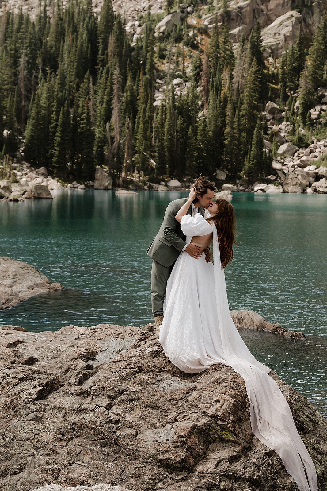 Rocky Mountain National Park elopement couple holding each other lakeside with massive rocky peaks and alpine forest behind them