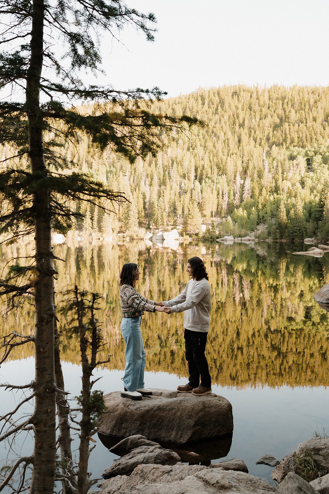 Couple holding hands on a boulder at Bear Lake RMNP with golden tree reflections in the still water