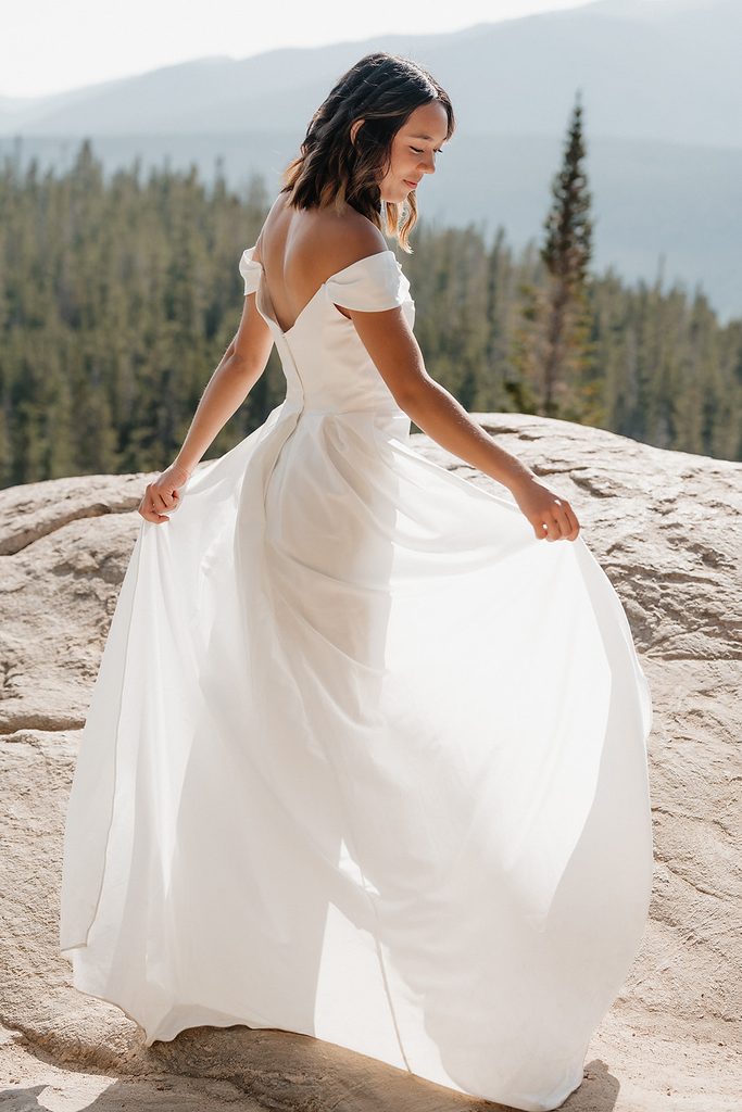 Bride in flowing white elopement gown twirling on rock ledge above Colorado pine forest and mountain range