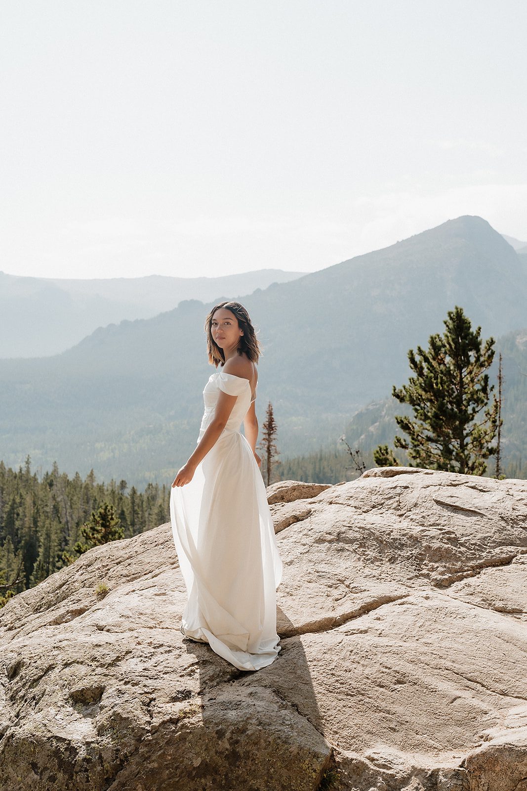 Bride in off-shoulder white gown standing on rocky overlook with Colorado peaks layered behind her